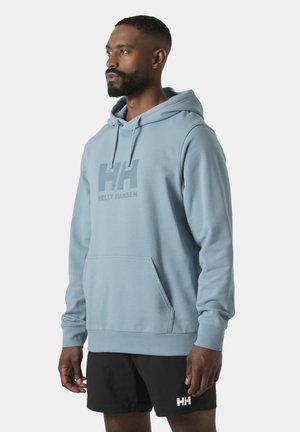 Hombre de pie, vistiendo una sudadera con capucha azul claro de Helly Hansen y pantalones cortos negros de Helly Hansen, mirando hacia la izquierda sobre un fondo claro y liso.