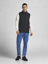 Zwarte gewatteerde vest over een witte langemouwen shirt, gecombineerd met lichtblauwe jeans en zwarte schoenen, met een rits aan de voorkant en een hoge kraag.