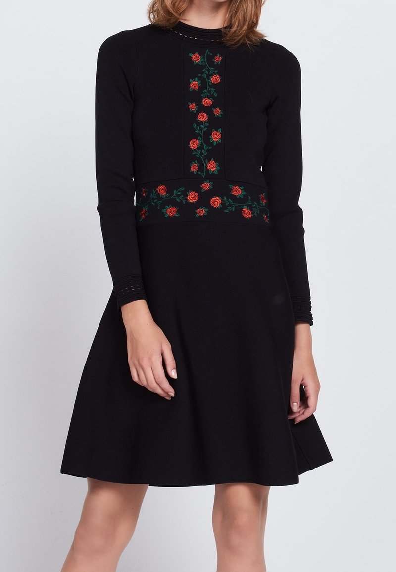 Femme portant une robe noire à manches longues avec une broderie de rose rouge sur la poitrine et la taille, debout devant un fond uni.