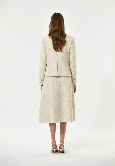 Costume texturé crème avec une veste courte et une jupe longueur genoux. Présente une finition lisse, des bords légèrement effilochés et des talons beiges assortis.