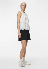 Haut sans manches en dentelle blanche à œillets avec un ourlet festonné, associé à un short noir et des baskets argentées ; le modèle se tient contre un fond uni.