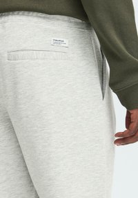 Pantalones de chándal gris claro hechos de una tela suave, con un bolsillo trasero que lleva una etiqueta y un ajuste holgado. La cinturilla es elástica para mayor comodidad.