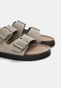Beige ruskindsandaler med to justerbare stropper, metalliske spænder og en sort gummisål med en tekstureret overflade.