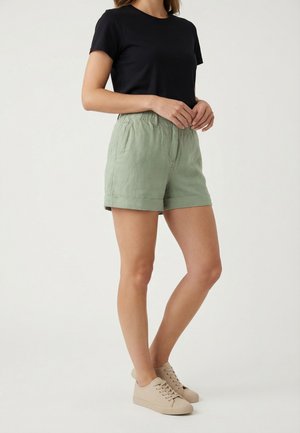 Mujer de pie con camiseta negra de manga corta, pantalones cortos verde claro con dobladillo enrollado y zapatillas beige sobre un fondo claro y liso.