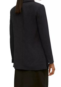 Blazer noir en tissu lisse et mat. Col cranté, manches longues et coupe cintrée. Détail de couture au dos visible.