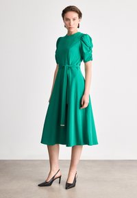 Πράσινο φόρεμα με φουσκωτά κοντά μανίκια, εφαρμοστό κορμό και φαρδιά φούστα. Περιλαμβάνει ζώνη στη μέση που δένει. Φοριέται με μαύρες κλειστές ψηλοτάκουνες εσπαντρίγιες.
