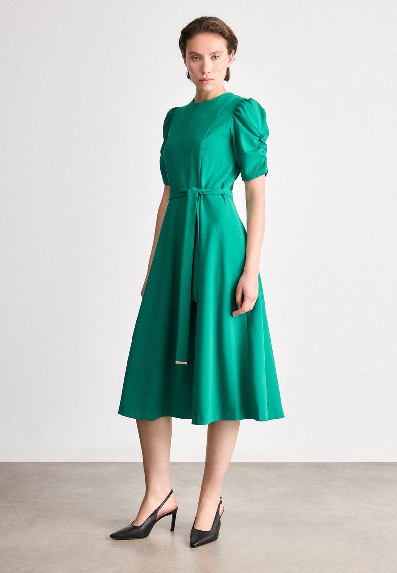 Πράσινο φόρεμα με φουσκωτά κοντά μανίκια, εφαρμοστό κορμό και φαρδιά φούστα. Περιλαμβάνει ζώνη στη μέση που δένει. Φοριέται με μαύρες κλειστές ψηλοτάκουνες εσπαντρίγιες.