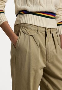 Person som bär beiga högmidjade veckade byxor med en knäppt ficklock och en krämfärgad tröja med färgglada ränder vid midjan och cuffs.