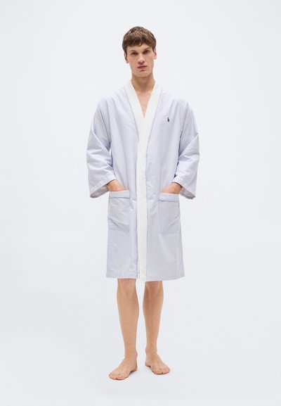 Νεαρός άνδρας που στέκεται ξυπόλητος, φοράει μπλε ανοιχτόριγμη ρόμπα μέχρι το γόνατο με λευκό περίγραμμα και μπροστινές τσέπες.