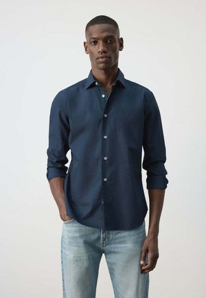 Hombre con una camisa azul oscuro abotonada con las mangas remangadas y vaqueros azul claro frente a un fondo claro y liso.