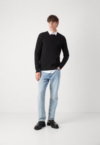 Zwarte trui met V-hals, gedragen over een witte overhemd met kraag, gecombineerd met lichtblauwe spijkerbroek en zwarte leren schoenen, staand op een witte achtergrond.