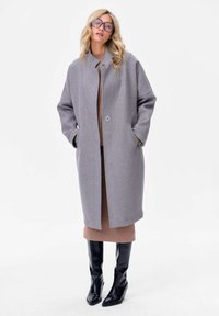 Manteau en mélange de laine gris clair avec col montant, fermeture à un bouton et poches latérales. Porté sur une robe marron et des bottes noires jusqu'au genou.