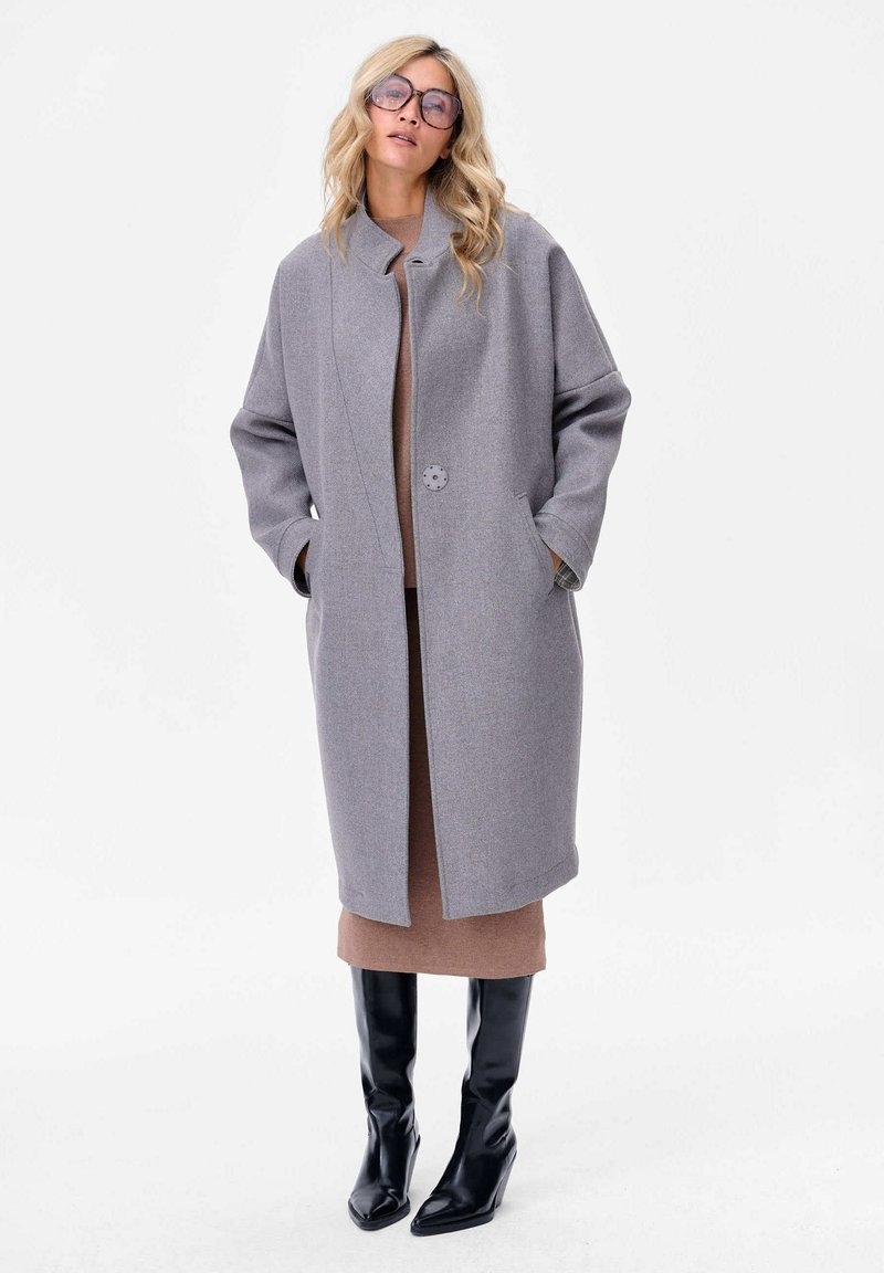 Manteau en mélange de laine gris clair avec col montant, fermeture à un bouton et poches latérales. Porté sur une robe marron et des bottes noires jusqu'au genou.