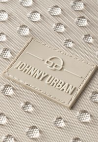 Superficie di una borsa in tessuto beige con accenti circolari in rilievo e una toppa logo in rilievo di colore chiaro con scritto "JOHNNY URBAN."