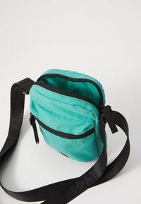 Borsa a tracolla teal con chiusura lampo, dotata di una tracolla nera impreziosita da testo in rilievo. Texture del tessuto liscia e forma rettangolare compatta.