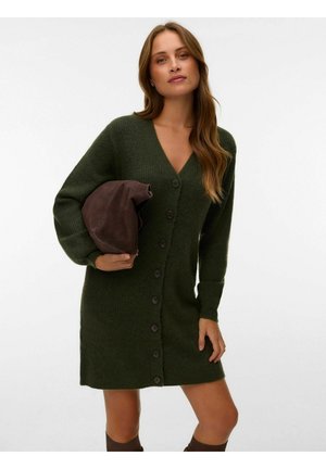 Donna che indossa un vestito a maglia verde scuro con bottoni che tiene una borsa piegata in camoscio marrone scuro su uno sfondo semplice.