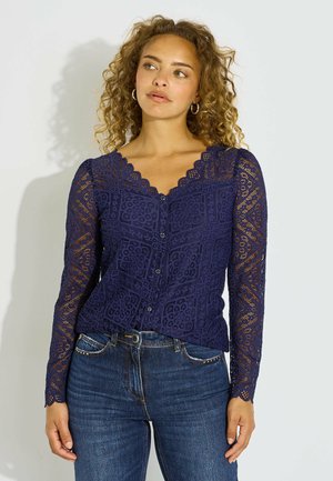 Mujer con cabello rizado que lleva una blusa azul marino de encaje con mangas largas y vaqueros azules, mirando ligeramente hacia un lado sobre un fondo liso.
