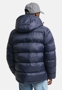 Marineblå pufferjakke med hette, med en quilted design og blank finish, parret med casual lyseblå jeans og en grå caps.