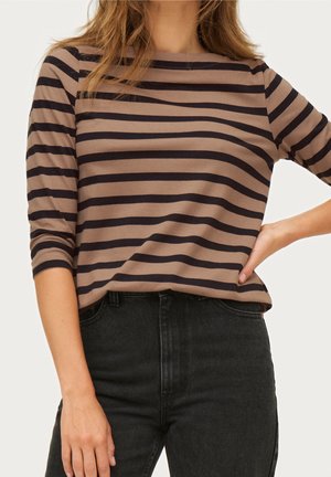 Frau trägt ein braun-schwarz horizontal gestreiftes Shirt mit dreiviertel Ärmeln und hoch taillierte schwarze Jeans, Hand in Hüfthöhe.