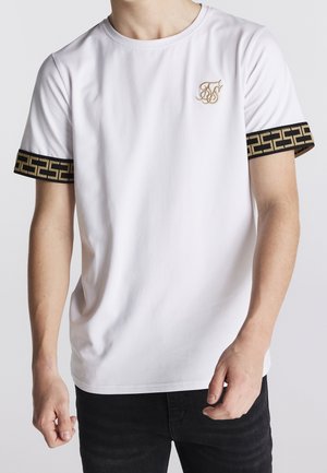Mand iført hvid kortærmet skjorte med sort og guld geometrisk trim på ærmerne og lille gylden broderet logo på brystet.