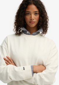 Sudadera color crema con un corte relajado y puños acanalados, superpuesta a una camisa de cuello a rayas con rayas blancas y marinas.
