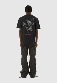 Sort oversized t-shirt med hvide sommerfugle-grafikker og orange tekst på ryggen, parret med mørke cargo bukser.