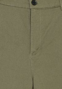 Lähikuva oliivinvihreistä housuista, joissa näkyvät nappi, vetoketju ja etuvyötärön ompelut.