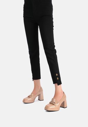 Damskie nogi w czarnych dopasowanych spodniach z złotymi guzikami przy kostkach oraz w beżowych lakierowanych loafersach na słupku z detalami w formie łańcucha.