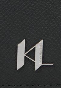 Juodas tekstūruotas medžiagos paviršius su sidabriniu metaliniu logo "KL", kuris turi kampuotus dizaino elementus ir įrengtas prekės pavadinimas.