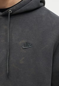 Κοντινό πλάνο σκούρου γκρι φούτερ Nike με κεντημένο λογότυπο στο στήθος και κορδόνι που κρέμεται από τη κουκούλα.