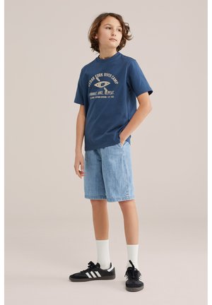 Fiú zsebre tett kézzel, sötétkék mintás pólót, világos farmershortot, fehér zoknit és fekete sportcipőt visel semleges háttér előtt.