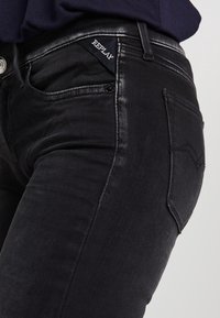 Jeans de denim negro con un diseño ajustado, ligero brillo y una etiqueta de logo en el lateral. Incluye bolsillos frontales y acentos de costura distintivos.