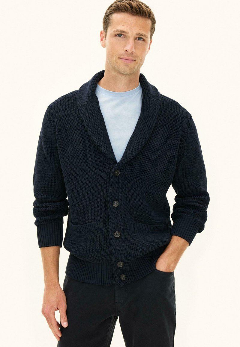 Uomo che indossa un cardigan blu navy con collo sciallato abbottonato sopra una camicia azzurra, in piedi con una mano in tasca davanti a uno sfondo semplice.