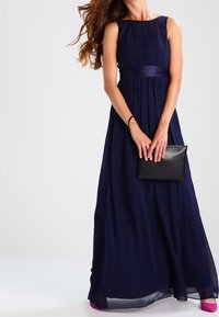 Abito maxi blu navy con corpino plissettato, design senza maniche e cintura in satin. Abbinato a una pochette nera e tacchi rosa.