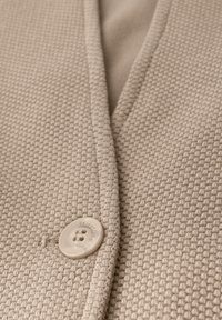 Tissu beige texturé avec un motif tissé, présentant une fermeture à bouton proéminente. Le col a une forme en V subtile et des détails de couture soignés.