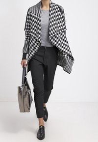 Černobílý vzorovaný cardigan s volným předním dílem, nošený přes šedé tričko, v kombinaci s černými slim-fit kalhotami a černými botami, nese kovovou tašku.