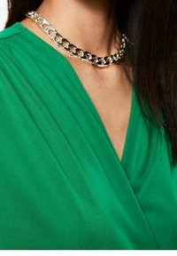 Blusa de tela verde con diseño de escote en forma de V y pliegues. Un collar de cadena de plata gruesa reposa alrededor del cuello, resaltando el escote.