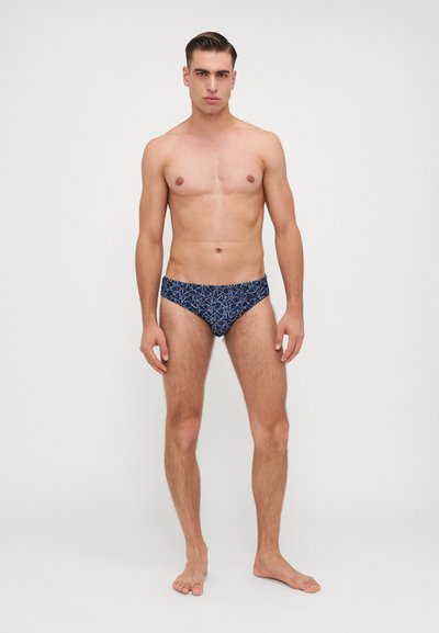 Marine zwemslips met een geometrisch wit patroon, aansluitend ontwerp, gemaakt van zachte stof. Model staat blootsvoets, met een neutrale achtergrond.