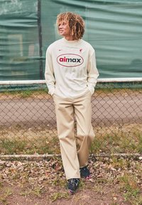 Camicia a maniche lunghe color crema con logo rosso e nero, pantaloni beige a gamba larga e sneaker nere con accenti rosa, in piedi vicino a una recinzione.