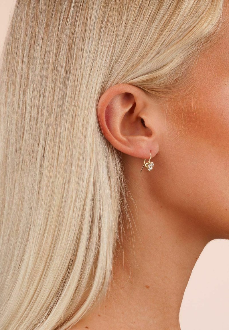 Gold earrings with a floral design featuring a clear gemstone, suspended by a thin hook, worn on a light-blonde hairstyle.