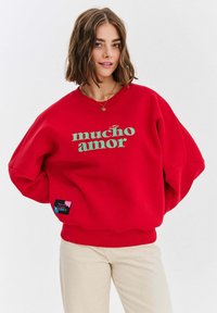 Czerwona bluza z okrągłym dekoltem z napisem "mucho amor" w kolorze zielonym, z metką w lewym dolnym rogu z różowymi i niebieskimi akcentami. Miękki materiał, luźny krój.