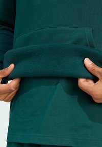 Tmavý teal sveter s mäkkým, prehnutým lemom a prednou kapsou. Materiál vyzerá hladko a elasticky, čím zvýrazňuje voľný dizajn.