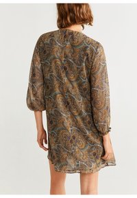 Femme portant une robe ample avec un motif paisley marron, bleu et vert, se tenant de dos face à la caméra, les bras détendus le long du corps.