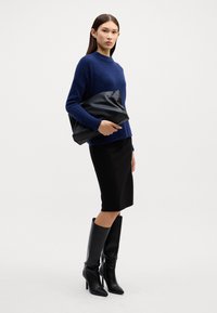 Dunkelblauer Strickpullover, schwarze figurbetonte Rock, schwarze kniehohe Stilettos, hält eine schwarze Lederhandtasche, minimalistisches Design, glatte Texturen.