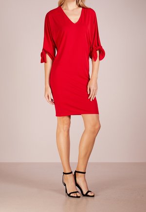 Femme portant une robe rouge mi-longue à col en V et manches trois-quarts nouées en nœuds, associée à des sandales noires à talons hauts.