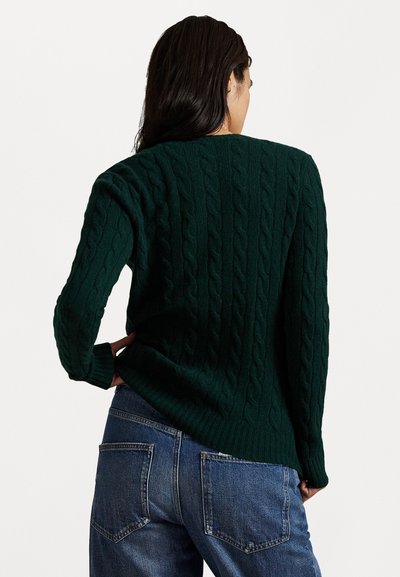 Groene kabelbrei-sweater met een textuurpatroon, lange mouwen en een geribbelde zoom, gecombineerd met een blauwe spijkerbroek. Focus op de achterkant.