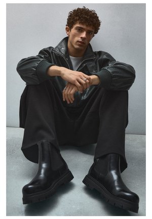 Jeune homme aux cheveux bouclés assis par terre portant une veste en cuir noire, un pantalon large noir, des bottes noires et des bagues en argent.
