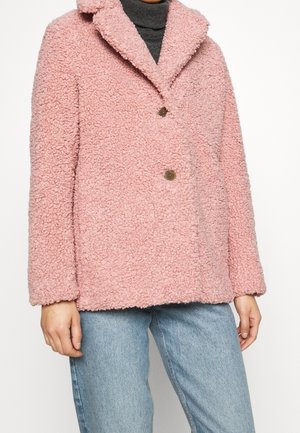 Femme portant un manteau en laine texturé rose clair avec deux boutons, un col roulé gris foncé et un jean bleu clair, vue des épaules aux cuisses.