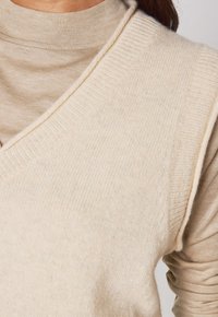 Gilet in maglia beige con scollo a V e bordi a costine, indossato sopra una maglietta a maniche lunghe color nocciola chiaro. Tessuto morbido e look a strati.