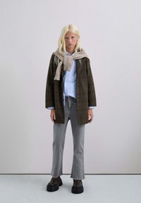 Jongeman met lange blonde haren, gekleed in een bruin geruite jas, een blauwe shirt, grijze versleten jeans, stevige zwarte laarzen en een beige trui op zijn schouders.